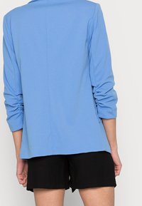 Vaaleansininen bleiseri, jossa on sileä pinta, rento leikkaus ja kolmen neljänneksen hihat. Takana on yksinkertainen ja siisti muotoilu. Yhdistetty mustiin shortseihin.