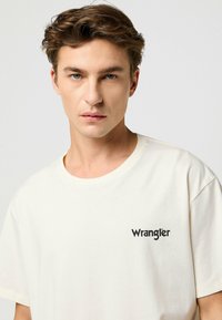 Krémové bavlnené tričko s okrúhlym výstrihom a krátkymi rukávmi, s čiernym logom "Wrangler" na ľavej hrudi.