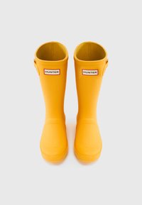 Κίτρινα λαστιχένια μπότες βροχής με λεία επιφάνεια, ίσια πλαϊνά και ετικ�έτες με το λογότυπο. Στρογγυλεμένες μύτες και διακοσμητικό αγκράφα στο εξωτερικό πλευρό.