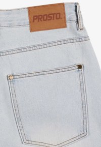 Jasnoniebieska tylna kieszeń dżinsów z denimu z podwójnym przeszyciem i brązową skórzaną metką z napisem "PROSTO." nad pasem.