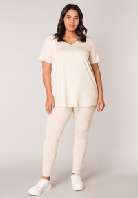 Beige T-shirt met korte mouwen en V-hals, gecombineerd met beige strakke legging, beide gemaakt van zachte, rekbare stof. Gecombineerd met witte casual sneakers.
