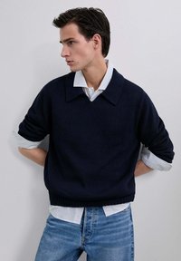 Suéter azul marino con textura de canalé y cuello ancho, superpuesto a una camisa a rayas azul claro. Combinado con jeans de mezclilla azul claro.