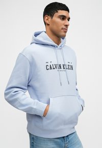 Svetlomodrá mikina s kapucňou, so šnúrkou, prednou kapsou a čiernym logom "CALVIN KLEIN". Mäkká látka, voľný strih. Neformálny dizajn a štýl.