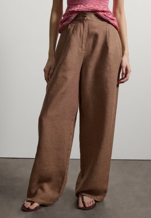 Frau trägt braune Leinenhose mit hoher Taille und weitem Bein, kombiniert mit braunen Flip-Flops und einem kurzen rosa Stricktop, steht auf grauem Boden.