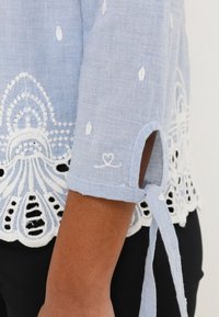 Lyseblå hørbluse med blonderkant, broderede detaljer og bånd ved ærmekanterne. Stoffet har en struktureret overflade med hvide accenter.