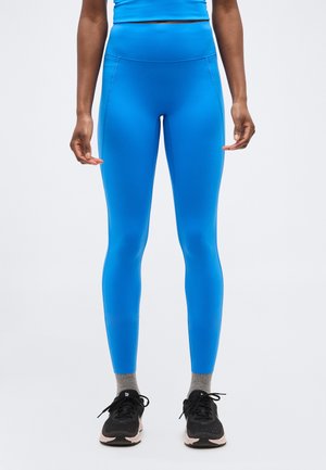 Person iført lyseblå højtaljede leggings, grå sokker og sorte sportssko, stående mod en ensfarvet hvid baggrund.