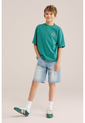 Garçon debout, une main dans la poche, portant une chemise à manches courtes bleu sarcelle, un short en denim clair, des chaussettes blanches et des baskets vertes sur un fond beige.