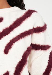 Krém színű pulóver texturált bordó szőrös csíkokkal, kerek nyakkal és kötött mintával. Puha anyag, enyhén bő fazonban.