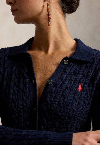 Nő tengerészkék kábelmintás polóingben, piros hímzett logóval és hosszú gyöngyös fülbevalóval, balra néz, haját kontyba fogta.