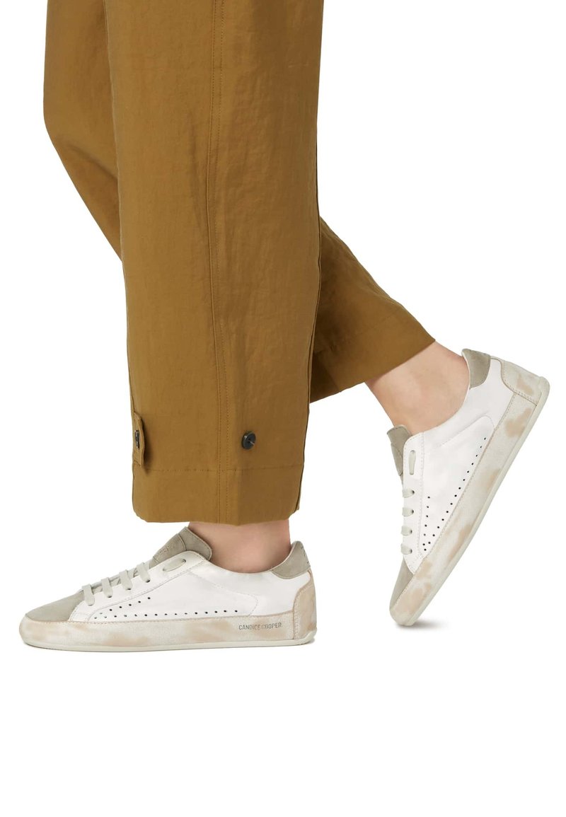 Weiße Sneaker mit beigen Wildlederelementen, durchbrochener Seitenverarbeitung und einer weichen, strukturierten Sohle. Getragen zu lockeren braunen Hosen.