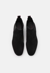 Paire de chaussures noires en suède à enfiler avec des languettes rouges, sur fond blanc, photographiées vue de dessus.