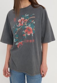 Szary, oversize t-shirt z bawełny, ozdobiony kwiatowym wzorem w różowym i zielonym kolorze z czerwonym napisem, krótki rękaw i okrągły dekolt.