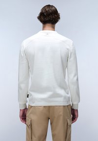 Pull over blanc avec manches longues, tissu texturé, poignets et col côtelés. Étiquette Woodin sur la hanche gauche. Porté avec un pantalon cargo beige.