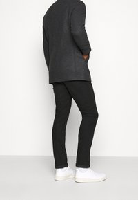 Uomo che indossa un blazer grigio scuro, jeans neri e sneakers bianche, in piedi contro uno sfondo bianco uniforme, con le mani nelle tasche.