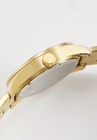 Guldarmbåndsur set fra siden, der viser kransen og en del af metalarmbåndet på en hvid baggrund.