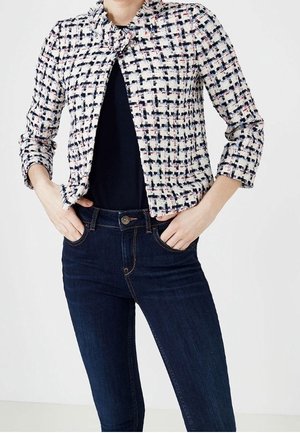 Femme portant une veste raccourcie noire, blanche et rose à motifs sur un haut noir avec un jean taille haute bleu foncé, mains dans les poches.