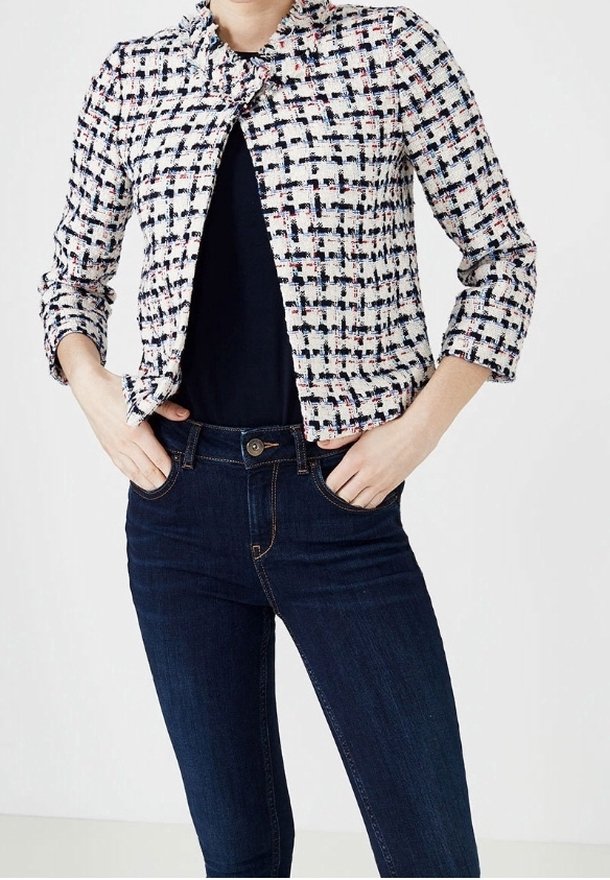 Femme portant une veste raccourcie noire, blanche et rose à motifs sur un haut noir avec un jean taille haute bleu foncé, mains dans les poches.