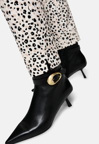 Schwarze Leder-Ankle-Boots mit spitzer Zehenform, ausgestattet mit einem goldenen, runden Akzent an einem schwarzen Riemen. Getragen zu Hosen im Leopardenmuster.