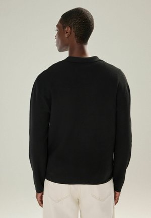 Sweater en tricot noir avec col, affichant une coupe décontractée et des manches longues, vu de dos, associé à un pantalon de couleur claire.