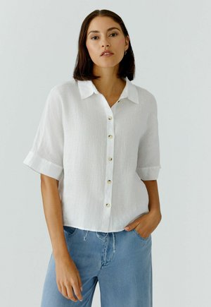 Frau mit kurzen dunklen Haaren, die ein weißes Hemd mit Knöpfen und hellblaue Jeans trägt, steht mit einer Hand in der Tasche vor einem einfachen Hintergrund.