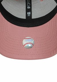 Gorra de béisbol con cuerpo de tela rosa y visera negra. Presenta una pegatina circular del logo de la MLB en la parte frontal. Superficie texturizada, diseño estructurado.