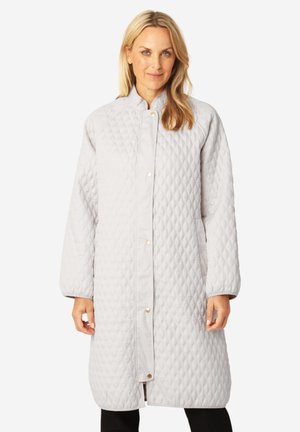 Donna con indosso un lungo cappotto trapuntato grigio chiaro con bottoni a pressione, in piedi davanti a uno sfondo bianco uniforme.