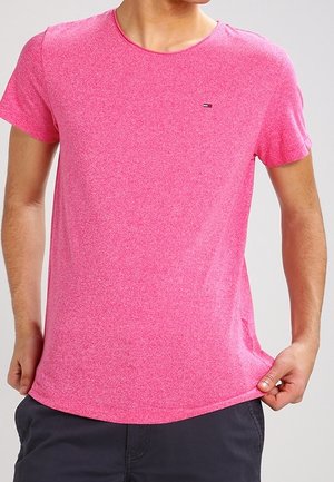 Man draagt felroze T-shirt met korte mouwen en houdt de zoom vast, gecombineerd met donkere korte broek, tegen een effen lichtgrijze achtergrond.