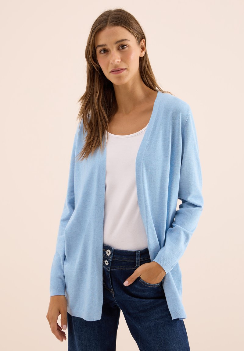 Jonge vrouw met lang bruin haar draagt een lichtblauwe cardigan over een wit topje en donkerblauwe spijkerbroek, staand tegen een effen beige achtergrond.