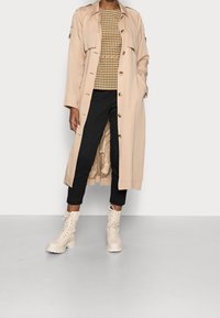 Trench coat beige con design doppiopetto, sopra una maglia a quadri. Pantaloni neri alla caviglia e stivali bianchi con lacci e suole spesse.
