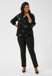Schwarze Bluse mit grünem Blumenmuster, V-Ausschnitt und Dreiviertelärmeln, kombiniert mit schwarzen, schmal geschnittenen Hosen und spitzen schwarzen Schuhen.
