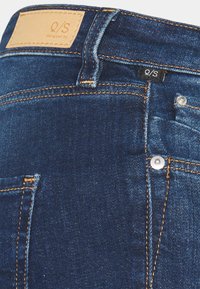 Dark blue denim jeans with orange stitching, a leather patch labeled "Q/S designed by," and a small black tag on the waistband.