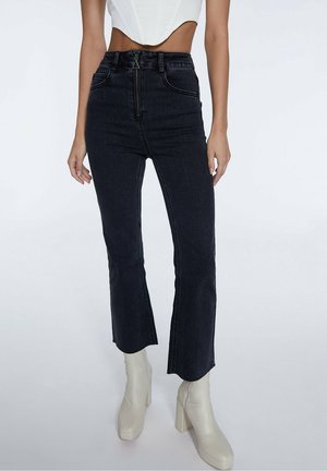 Jean flare en denim foncé taille haute avec fermeture éclair, dotés de poches avant et d'une texture lisse. Porté avec des bottes blanches à talons.