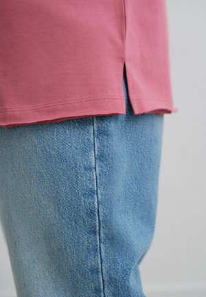 Chemise rose à manches courtes avec fentes sur les côtés, associée à un pantalon en denim bleu clair avec une texture et des détails de couture visibles.