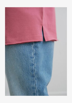 Chemise rose à manches courtes avec fentes sur les côtés, associée à un pantalon en denim bleu clair avec une texture et des détails de couture visibles.