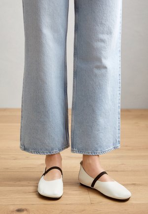 Białe płaskie buty z wycięciami, z czarnym paskiem wokół kostki. Noszone z jasnoniebieskimi szerokimi dżinsami na drewnianej podłodze.