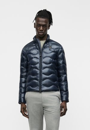 Muž s tmavou pokožkou a dredmi, oblečený v lesklom námorníckom prešívanom kabáte a svetlosivých nohaviciach, stojaci na jednoduchom svetlom pozadí.