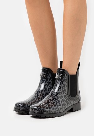 Bottes de pluie noires vernies à la cheville avec motif répété "MK" et boucle logo "MK" argentée, portées sur jambes nues sur un fond blanc uni.
