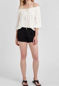 Žena v bílém off-shoulder topu, černých šortkách a černých sandálech, stojící na jednobarevném světle šedém pozadí.
