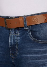 Brauner Leder Gürtel mit matter Oberfläche, schwarzer Metall-Schließe, kombiniert mit blauen Jeans aus Denim mit heller Naht und klassischem Fünf-Taschen-Design.