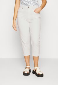 Jeans de cor clara, com comprimento até ao tornozelo e cintura ajustada, apresentando bolsos frontais e um tecido liso, ligeiramente texturizado. Combinados com sandálias brancas chunky.