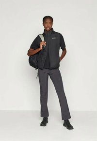 Černá zateplená vesta s horizontálním prošíváním, černé tričko, šedé kalhoty a černé boty. Model drží černý batoh a stojí proti neutrálnímu pozadí.