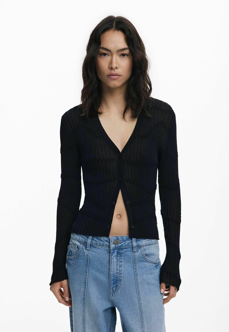 Zwarte geribbelde gebreide cardigan met een diepe V-hals en knoopsluiting, voorzien van textuurpatronen en lange mouwen, gecombineerd met lichtblauwe jeans.