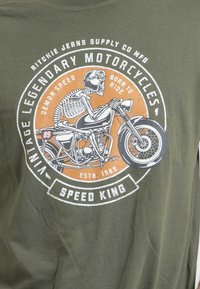 T-shirt vert olive avec un graphisme blanc et orange représentant un squelette cavalier sur une moto, entouré de texte circulaire et de détails.