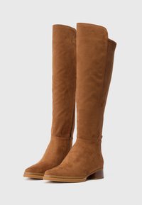 Bottes montantes en daim marron, à bout arrondi, avec un petit talon bloc et des coutures minimales. Texture lisse avec une finition légèrement glossy.