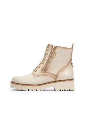 Botines de cuero beige con un cierre de cordones en la parte delantera, cremallera metálica en el lateral y adornos de tachuelas. Presentan una suela blanca gruesa con bordes dentados.