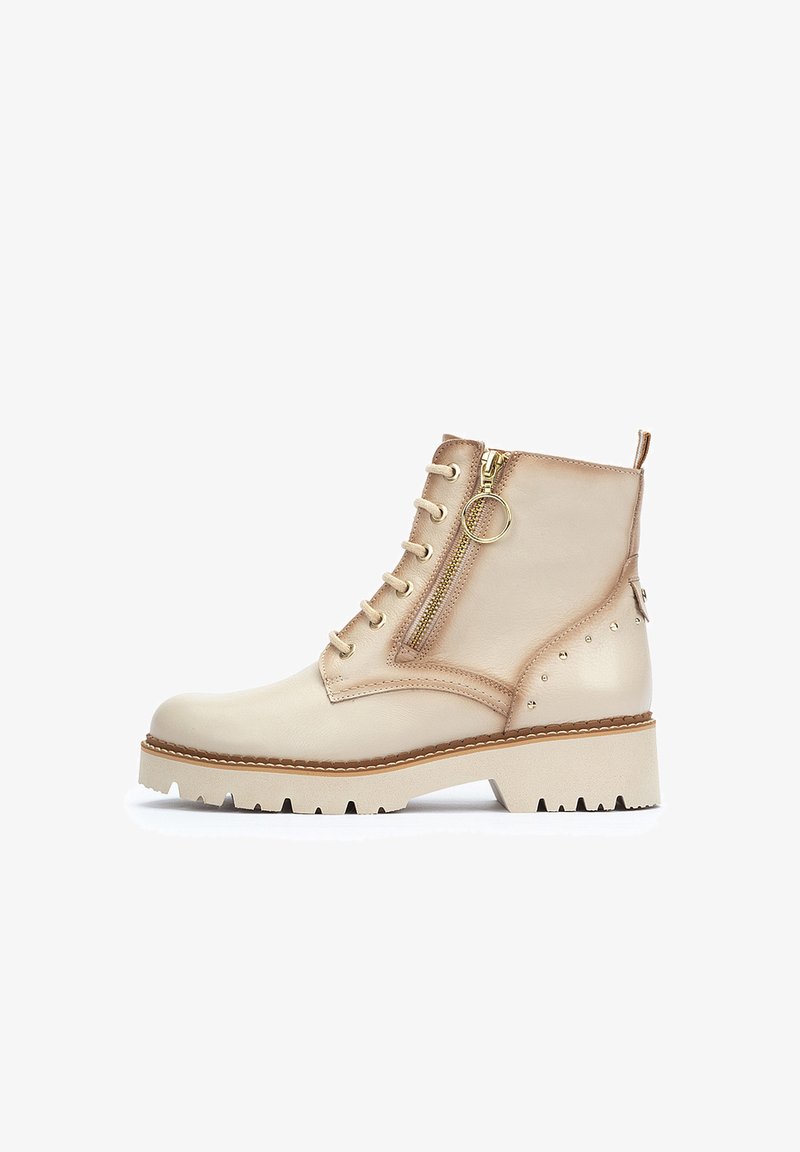 Botines de cuero beige con un cierre de cordones en la parte delantera, cremallera metálica en el lateral y adornos de tachuelas. Presentan una suela blanca gruesa con bordes dentados.