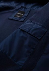 Marinblå jacka med en mjuk fleecekropp och släta nylonärmar, med en fyrkantig lapplucka och svart etikett inuti.