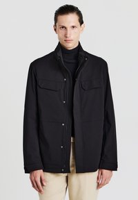 Giacca nera impermeabile con colletto alto, chiusura con bottoni e due tasche sul petto, caratterizzata da una texture liscia e maniche a taglio dritto.