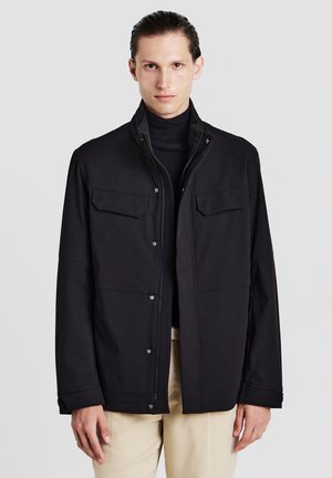 Veste noire imperméable avec un col haut, un front boutonné et deux poches poitrine, présentant une texture lisse et des manches droites.