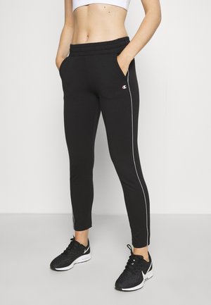 Osoba ubrana w czarne sportowe spodnie dresowe z białymi paskami po bokach, biały krótki top i czarne buty do biegania, stojąca z rękami w kieszeniach.